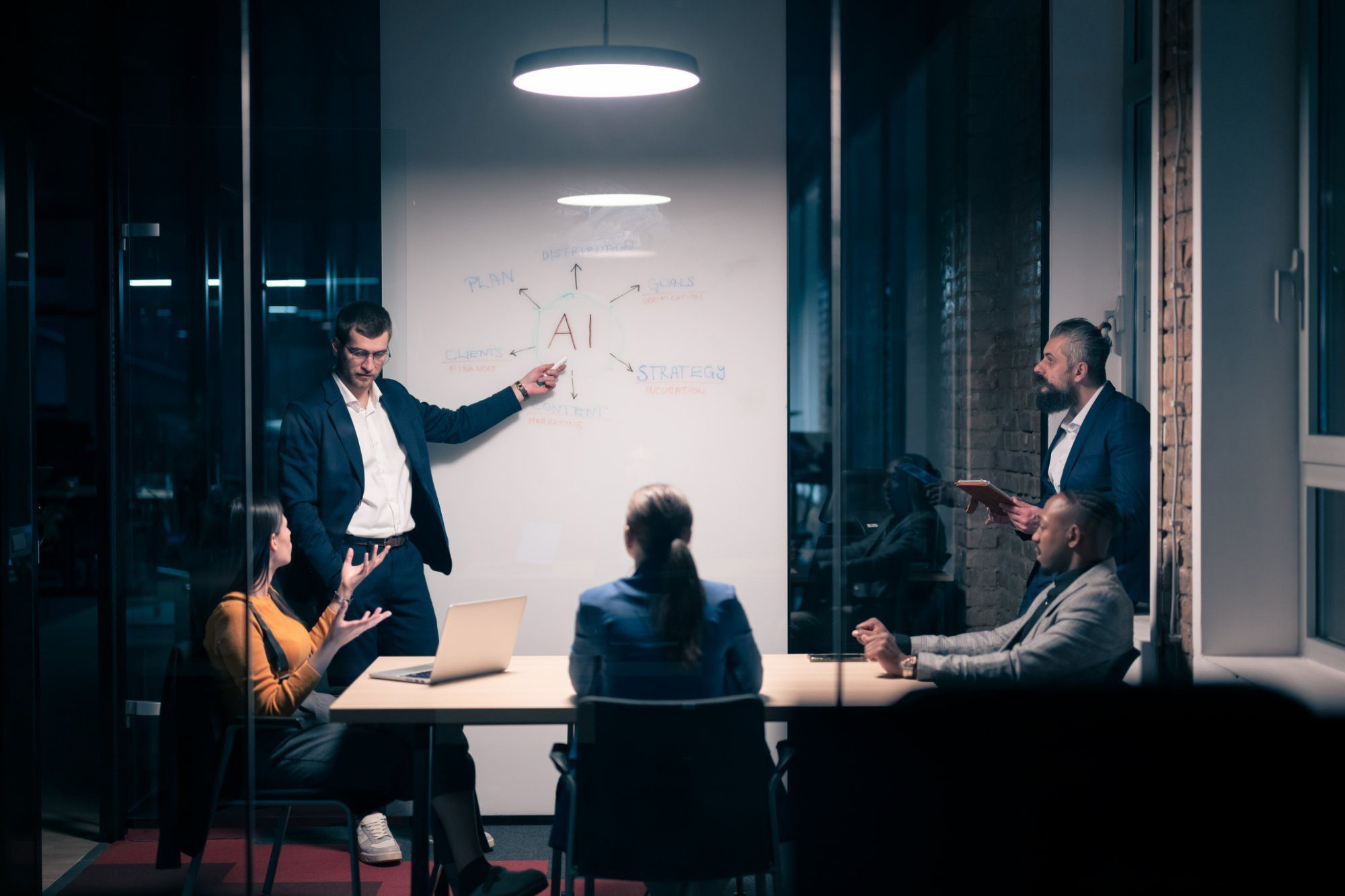 A marketing team meets in a conference room while a presenter points to a whiteboard diagram with AI at the center, connected to plan, distribution, goals, strategy, clients and content